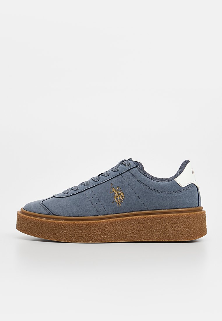Zapatilla de lona azul con suela texturizada marrón, acento blanco en el talón y emblema dorado. Cuenta con seis ojetes para los cordones y un diseño minimalista.