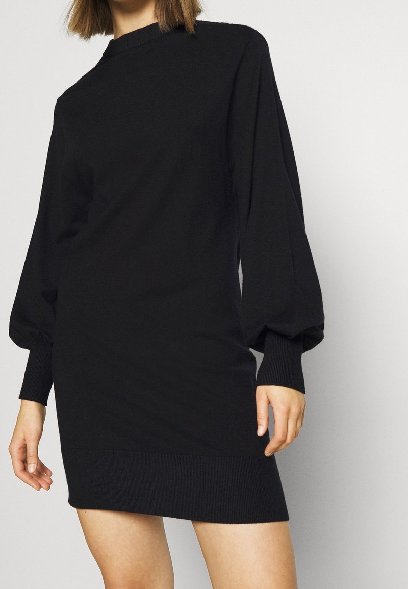 Femme portant une robe noire en maille à manches longues avec des manches bouffantes et des poignets côtelés, debout devant un fond uni.