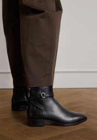 Stivali neri in pelle alla caviglia con punta rotonda, dettagli in cinturino metallico e tacco basso a blocco, abbinati a pantaloni marroni a sigaretta.