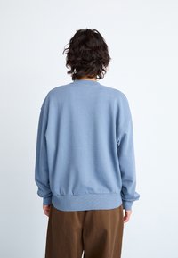 Blue sweatshirt with a crew neck, ribbed cuffs, and hem. Soft texture and relaxed fit, worn with brown pants. View from the back.