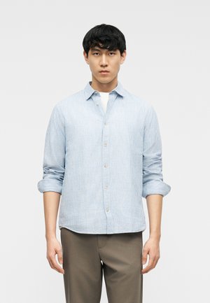 Man wearing light blue striped button-up shirt with sleeves rolled up, standing against plain white background.