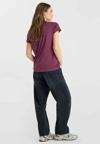 Vrouw met lang bruin haar, gekleed in een paarse T-shirt met korte mouwen, losse zwarte jeans en grijze sneakers, staande met haar handen in haar zakken.