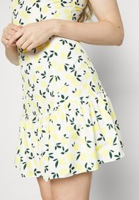 Robe blanche ornée d'un motif de citrons et de feuilles vertes, jupe évasée avec des détails froncés à la taille, longueur mi-cuisse. Texture de tissu lisse.