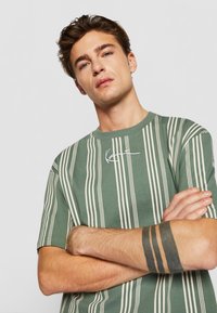Grönrandig t-shirt med krämfärgade vertikala ränder, rund halsringning och broderad logotyp. Tyget verkar mjukt med en avslappnad passform.