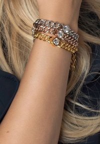 Drie kettingarmbanden in zilver, roségoud en goudtinten, met ronde heldere edelstenen. De kettingen zijn onderling verbonden met een soepele textuur.