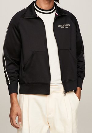 Homme portant une veste noire zippée avec des rayures blanches sur les manches et le texte "Hilfiger New York", associée à un pantalon et une chemise blancs.