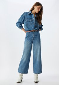 Camisa de mezclilla azul clara con dos bolsillos en el pecho, combinada con pantalones de mezclilla anchos de tiro alto en azul claro. Cinturón de cuero marrón y botas de tobillo blancas.