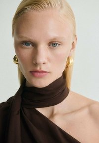 Un tessuto marrone si drappeggia attorno al collo; capelli biondi e lisci incorniciano il viso. Grandi orecchini d'oro mettono in risalto gli occhi blu e il tono di pelle chiaro.