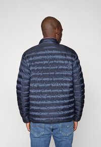 Marinegrønn pufferjakke med horisontalt quiltet mønster, høy krage og glatt tekstur. Bært over blå jeans, har en minimalistisk design.
