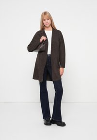 Cárdigan de lana marrón con un frente largo y abierto, combinado con jeans de mezclilla oscuros y de pierna acampanada, y botas negras hasta el tobillo. Lleva un pequeño bolso gris.