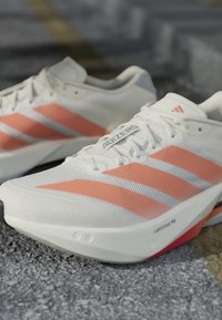Λευκά και ροζ αθλητικά παπούτσια Adidas με τρεις ρίγες και το λογότυπο "ADIZERO" στη διχτυωτή επάνω επιφάνεια, τοποθετημένα σε υφή εδαφικής επιφάνειας.