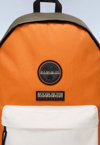 Orange ryggsäck med en mörkgrön topp, med en cirkulär logotyp-patch, en vit framlucka och ett texturerat material.