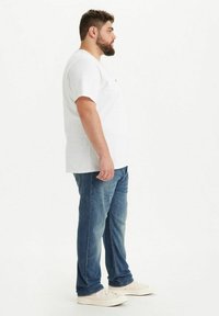 Valkoinen lyhythihainen t-paita pyöreällä kaula-aukolla, yhdistettynä sinisiin farkkuihin ja vaaleisiin lenkkareihin, näkymä sivusta.