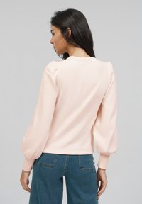 Lyse rosa genser med puffermer og rund hals. Mykt stoff med en jevn tekstur. Kombinert med blå denimbukser.
