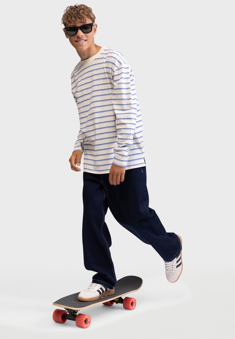 Long-sleeve white shirt with blue stripes, dark blue wide-leg pants, riding a skateboard with red wheels and wearing sunglasses.