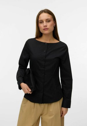 Kobieta z prostymi włosami, ubrana w czarną, zapinaną na guziki koszulę z długim rękawem oraz beżowe spodnie, trzymająca czarną kopertówkę na jednolitym tle.