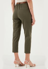Pantaloni croppati verde oliva con elastico in vita e tasca posteriore, abbinati a tacchi a strappo di colore chiaro. Texture liscia e vestibilità su misura.