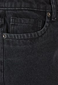 Tela de mezclilla negra con una textura suave, que presenta un bolsillo frontal y un acento de botón de metal. La costura visible aporta estructura al diseño.