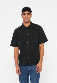 Črna kratka majica s kratkimi rokavi iz bombaža, z gumbom spredaj, koničastim ovratnikom in žepom na levi strani prsnega dela z znamkam.