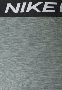 Vihreät urheiluleggingit, joissa on teksturoitu pinta ja musta vyötärönauha, jossa on valkoinen "NIKE"-logo. Kankaat näyttävät joustavilta ja hengittäviltä.