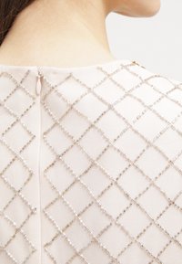 Beige plagg med ett diamantmönster av klara, rektangulära pärlor. Har en dragkedja i ryggen. Mjuk tygstruktur.