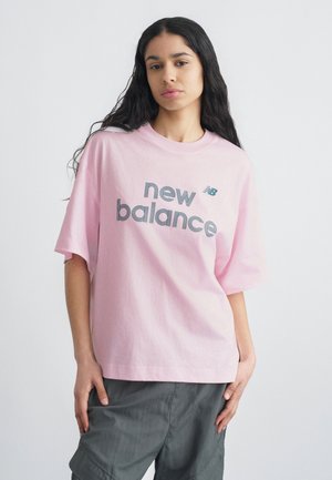 Donna con capelli lunghi e scuri che indossa una maglietta rosa larga della New Balance e pantaloni grigi, in piedi davanti a uno sfondo bianco semplice.