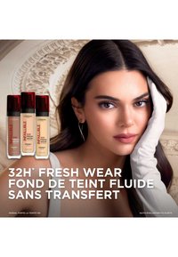 Bouteilles de fond de teint L'Oréal Infallible 32H Fresh Wear avec des bouchons rouges en différentes nuances, présentées aux côtés d'un modèle avec une peau lisse et claire portant des gants.