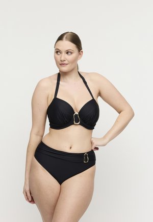 Vrouw poseert in een zwarte halterbikini met gouden gespsdetails, kijkt naar links tegen een effen lichte achtergrond.