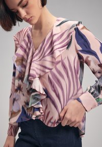 Blouse à motif floral en rose doux avec des volants le long de l'encolure et des manches ; présente un col en V et des manches longues avec des détails de poignet.