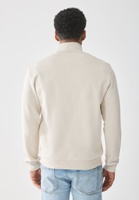 Sudadera beige con cuello alto, mangas largas y dobladillo acanalado. Confeccionada en una tela suave, con un acabado texturizado, llevada sobre unos jeans azules.