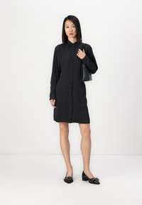 Robe chemise noire à boutons avec des manches longues, texture côtelée et longueur moyenne. Associée à des chaussures noires et un petit sac à main noir.
