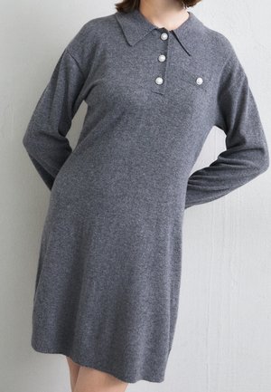 Kvinde iført en grå strikket kjole med lange ærmer, krave og fire perle-lignende knapper ned foran, stående mod en lys væg.