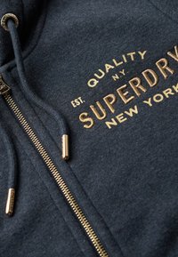 Sudadera con cremallera navy azul con logo bordado en dorado "SUPERDRY" y texto adicional. Presenta una cremallera dorada y detalles en cuerda. Textura suave.