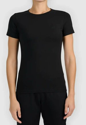 Kobieta ubrana w prosty czarny dopasowany t-shirt z krótkim rękawem oraz pasujące czarne spodnie, stojąca na jasnoszarym tle.