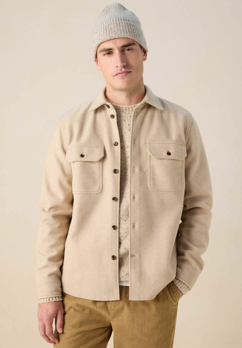 Veste en laine beige avec deux poches poitrine, boutons foncés et un ourlet arrondi. Portée par-dessus un pull léger et texturé ainsi qu'un pantalon en velours côtelé.