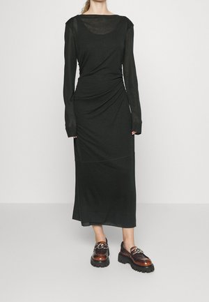 Kvinde iført en lang sort tætsiddende kjole med lange ærmer og brune chunky loafers med sølvkædedetalje.