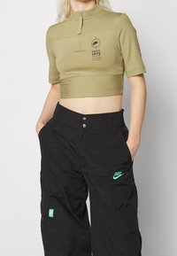 Olivově zelený zkrácený zipový top s krátkými rukávy a černé cargo kalhoty s zeleným logem Nike. Hladký materiál a uvolněný střih.