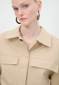 Camicia button-down beige con colletto, dotata di due tasche sul petto e una finitura liscia opaca. Il modello indossa grandi orecchini ad anello.