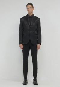 Completo nero testurizzato con rever in raso, abbinato a una camicia coordinata e pantaloni aderenti. Scarpe nere lucide completano il look.