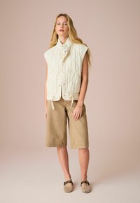 Gilet imbottito avorio con colletto alto, chiusura a bottoni snap e pantaloni corti in velluto a coste marrone chiaro. Indossato con scarpe beige a fantasia. Sfondo semplice.