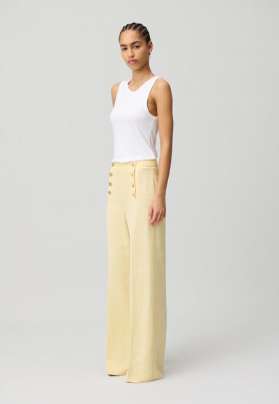 Un débardeur blanc sans manches associé à un pantalon jaune taille haute à jambes larges, agrémenté de cinq boutons décoratifs sur le devant.
