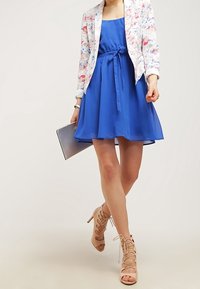 Femme marchant vêtue d'une robe bleue jusqu'aux genoux, d'un blazer à fleurs, de talons hauts nus à lacets, et tenant une pochette argentée.