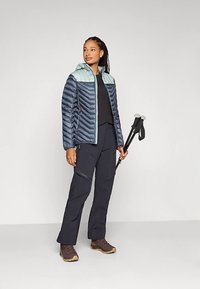 Woman in two-tone blue puffer jacket, black outdoor pants, and brown hiking boots holding black trekking poles, standing against a plain background.