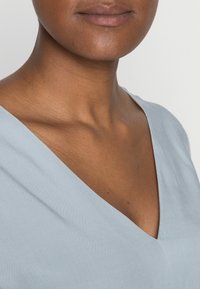 Nahaufnahme des Nackens und unteren Gesichts einer Person, die ein hellblaues V-Ausschnitt-Shirt vor einem schlichten Hintergrund trägt.