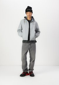 Lichtgrijze windbreakerjas met capuchon, zwart shirt eronder, grijze spijkerbroek en zwart met rode accenten sneakers. Zwarte beanie met tekst.