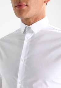 Man in een witte geknoopte dress shirt met een puntige kraag, weergegeven van kin tot borst tegen een effen achtergrond.
