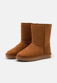 Hnědé semišové ankle boty s měkkou texturou, plochou gumovou podrážkou a jednoduchým designem. Švy podrobně vypracované podél švů.