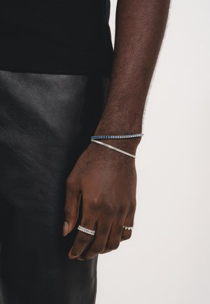 Silberner und blauer Armreif an einem Handgelenk, begleitet von mehreren Ringen. Der Armreif ist mit kleinen blauen Steinen verziert, und die Ringe haben klare geometrische Muster.