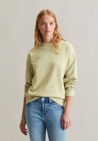 Lichtgroene sweatshirt met ronde hals en ribbelmanchetten, heeft subtiele branding in het midden. Draagt met blauwe spijkerbroek.