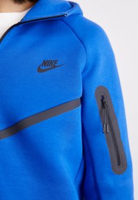 Manta azul da Nike com um logo preto, detalhes cinzentos e um bolso com fecho no braço. Feita de tecido texturizado com um design ajustado.